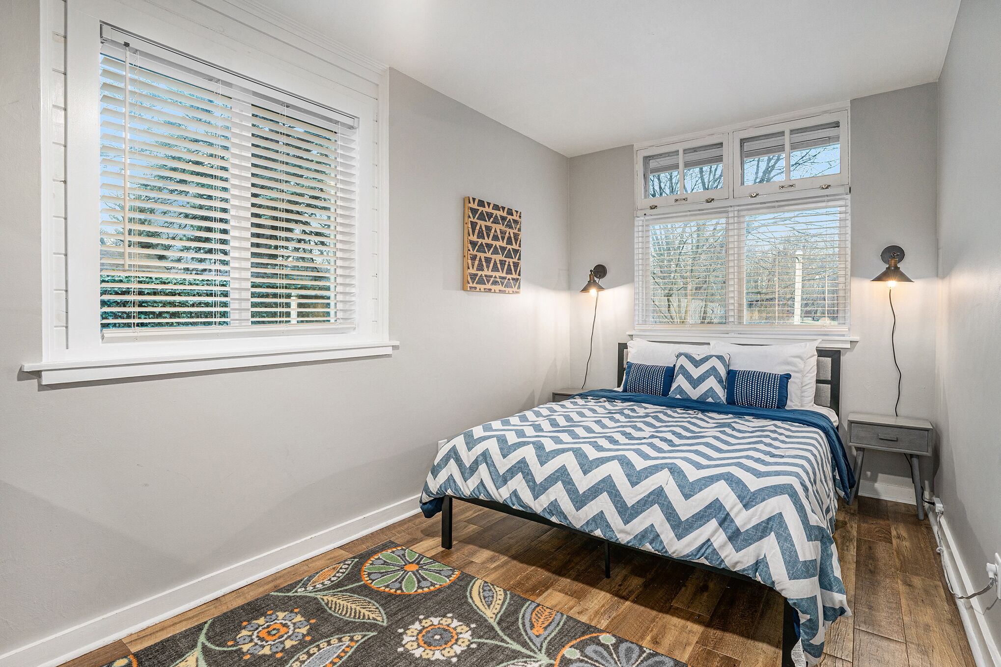 A bedroom with a bed, two bedside lamps, a window with blinds, and a patterned rug.