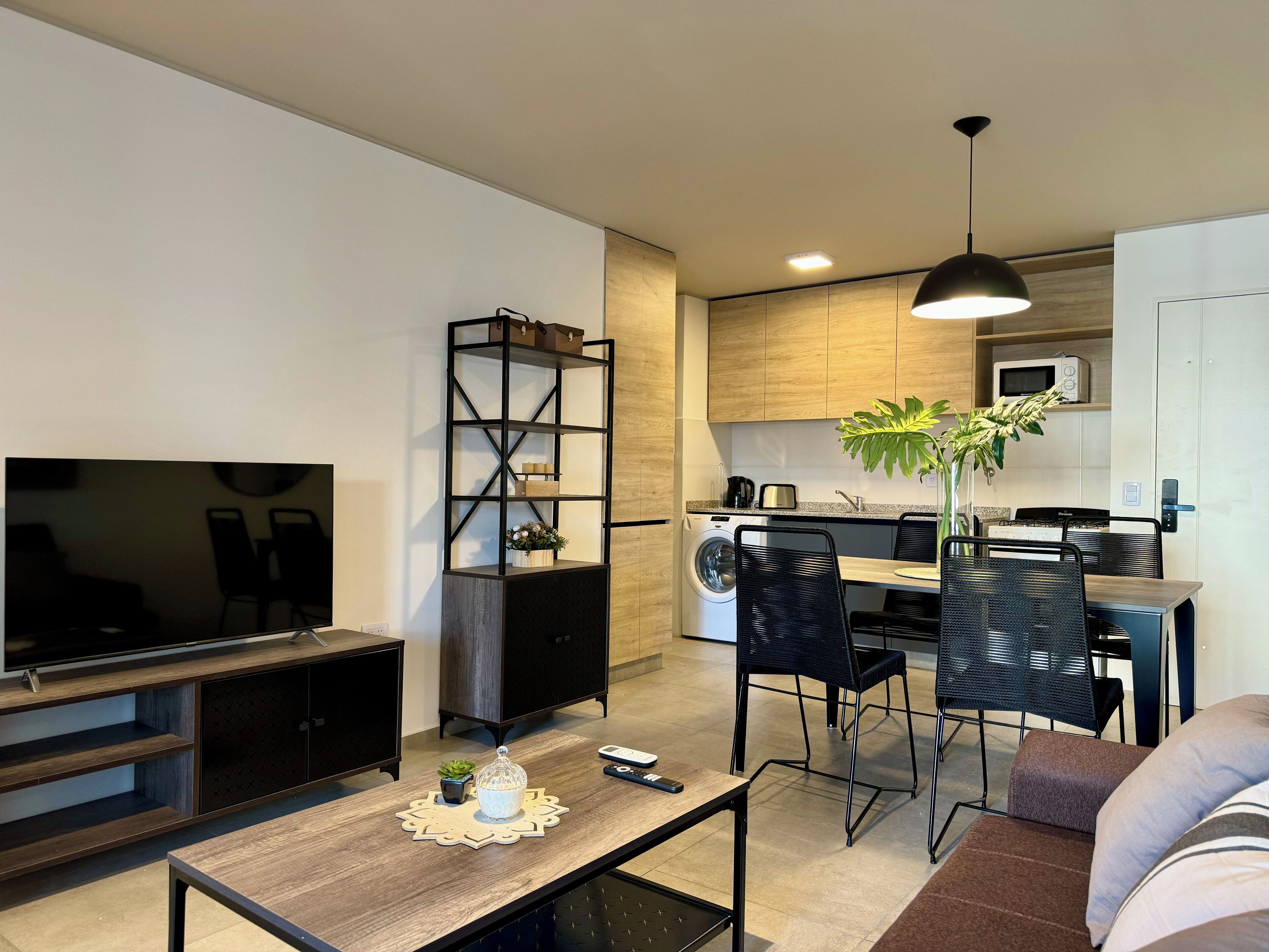 A modern living room with a TV, coffee table, and seating area. A kitchen with wooden cabinets and appliances is visible in the background.