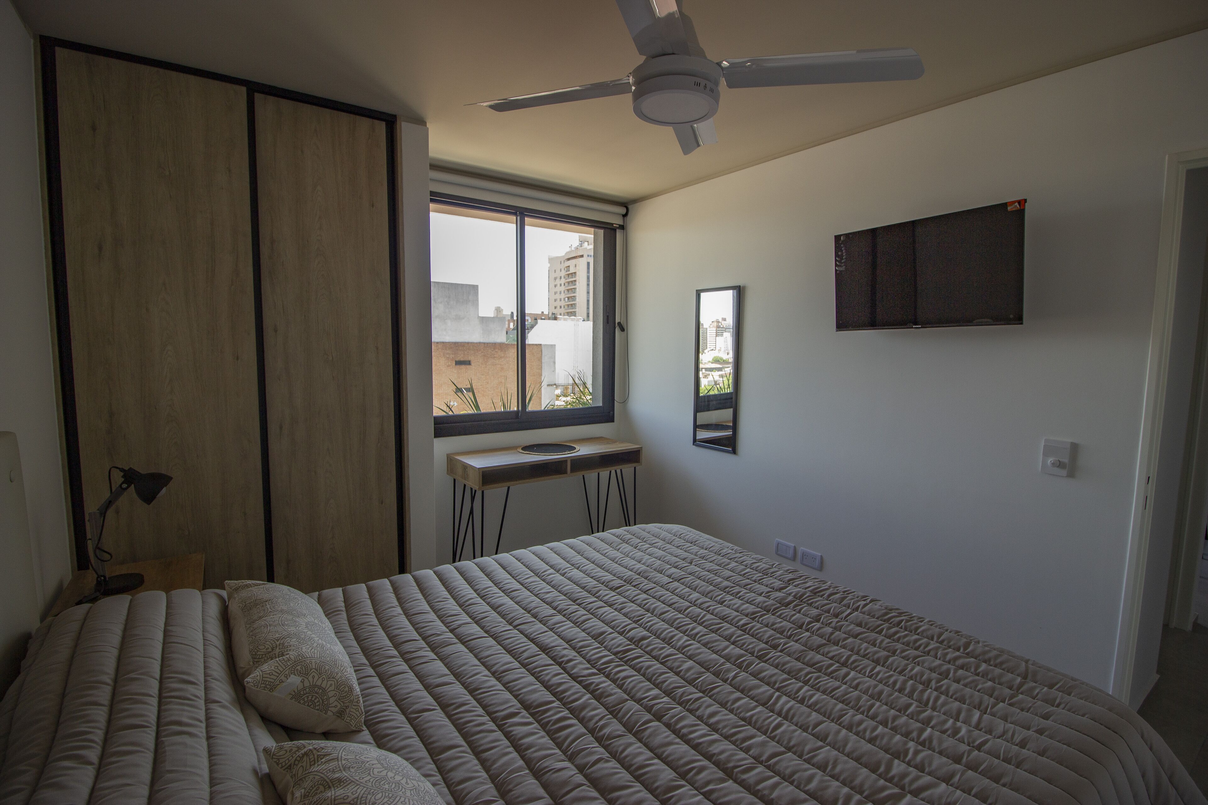A bedroom with a bed, a desk, a ceiling fan, and a window with a view of buildings.