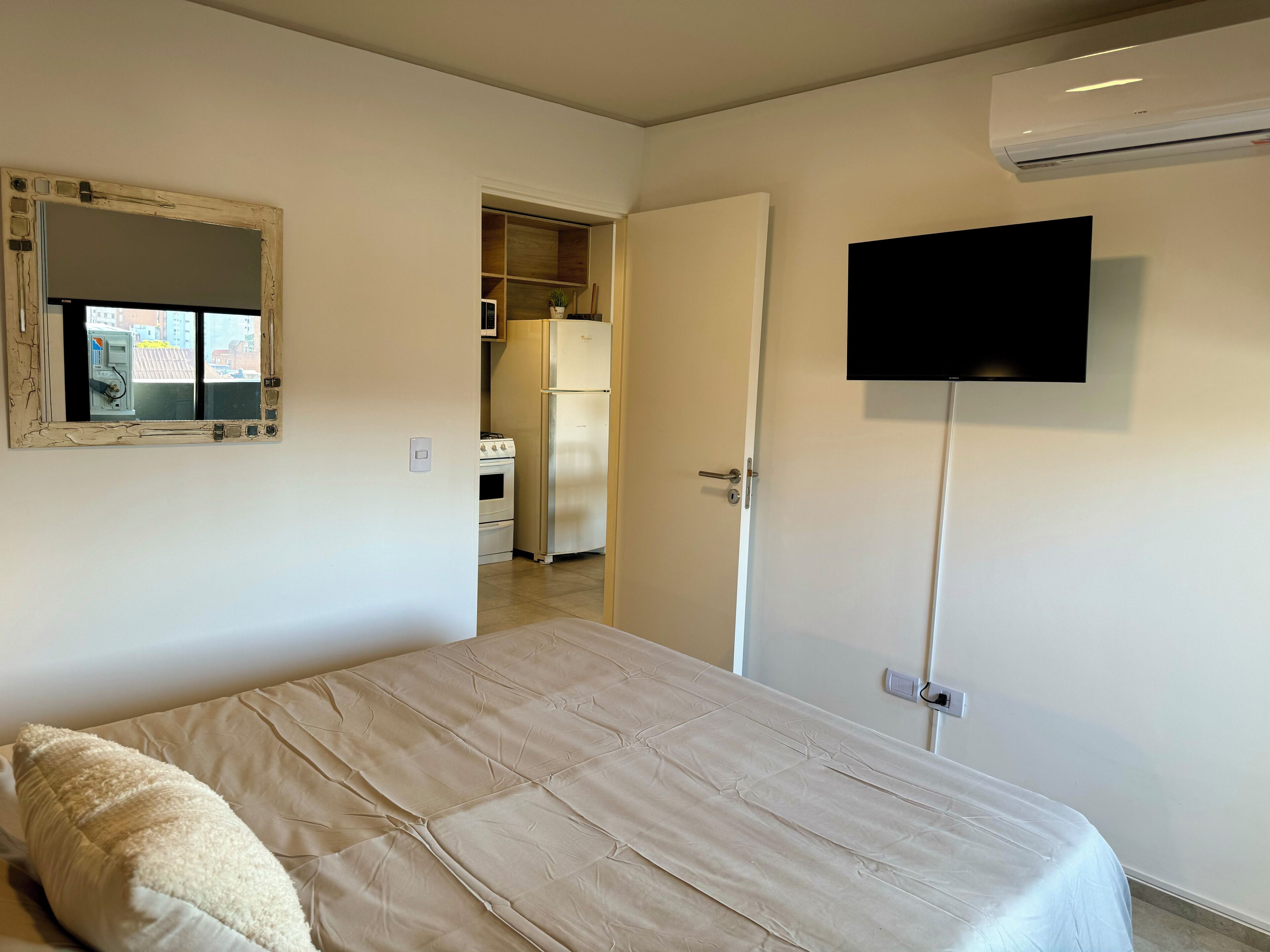 A bedroom with a bed, a television mounted on the wall, and a kitchen visible through an open door.