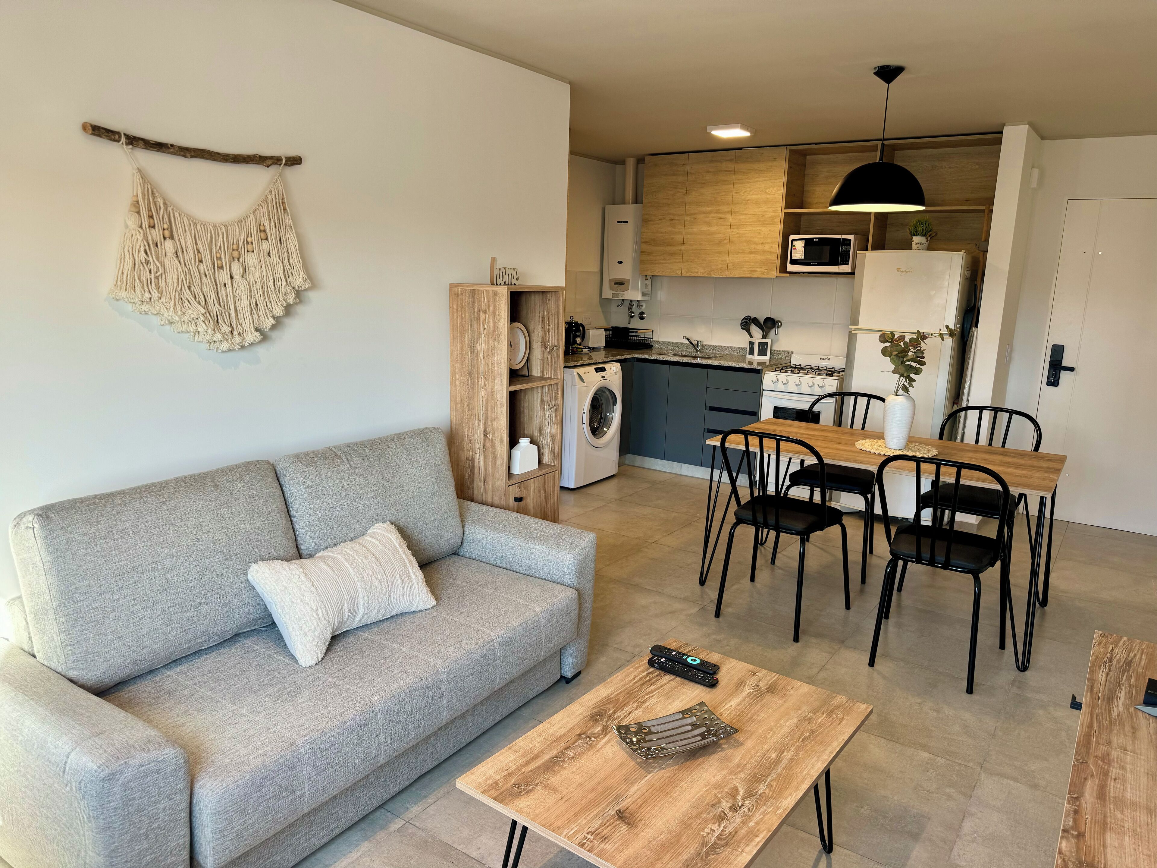 Un apartamento moderno con sofá gris, mesa de comedor de madera y sillas negras. Se ve una cocina con microondas, estufa y fregadero.