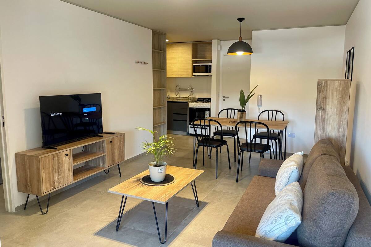 A modern living room with a flat-screen TV, a wooden coffee table with a plant, a grey sofa, and a dining area with a wooden table and chairs.