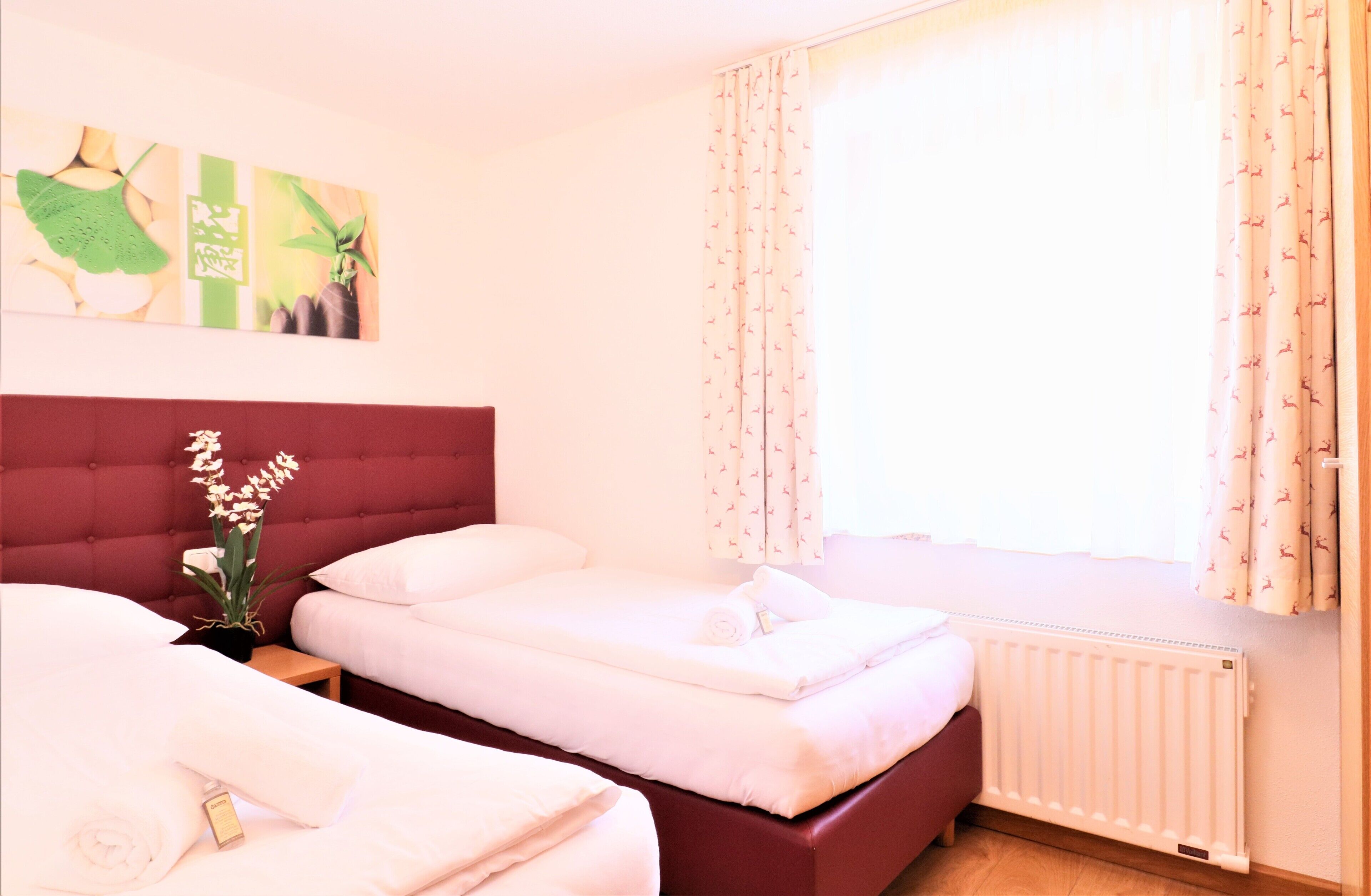 A hotel room with two beds, a red headboard, a window with patterned curtains, and a radiator.