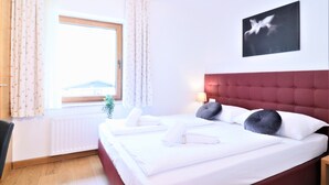 Una cama bien tendida con sábanas y almohadas blancas, un cabecero rojo, una ventana con cortinas y un cuadro colgado en la pared.