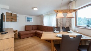 Una sala de estar moderna con una mesa de comedor de madera preparada para dos personas, un sofá de cuero marrón y un televisor sobre un mueble de madera.