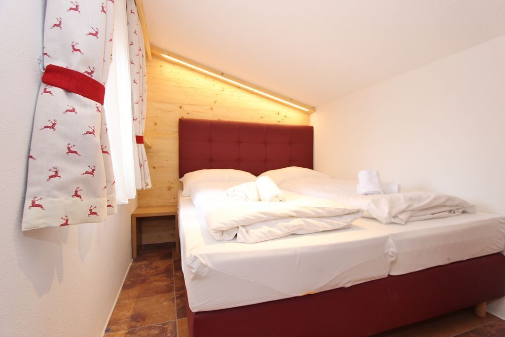 Une chambre à coucher avec une tête de lit rouge, du linge de lit blanc et un rideau orné d’un motif rouge.