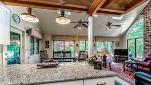 A spacious kitchen with granite countertops, white cabinets, and a large island. There are ceiling fans, a dining area with a red rug, and a living room with a leather couch and a TV.
