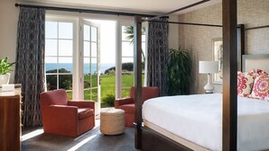 Ein Bett mit Baldachin und Blick auf den Ozean, ein Schreibtisch mit Lampe sowie zwei Sessel in einem Zimmer mit großen Fenstern.
