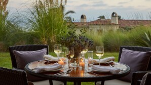 Une table de salle à manger ronde, dressée pour quatre personnes, avec des serviettes blanches, des verres à vin et des bougies, entourée de chaises en osier dotées de coussins, le tout se détachant sur un fond de hautes herbes et d’un bâtiment.