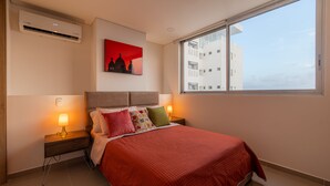Moderni hotellihuone, jossa on suuri sänky, punainen peitto, kaksi yöpöytävalaisinta, yöpöytä, seinälle asennettu ilmastointilaite ja suuri ikkuna, josta avautuu näkymä kaupungin ylle.
