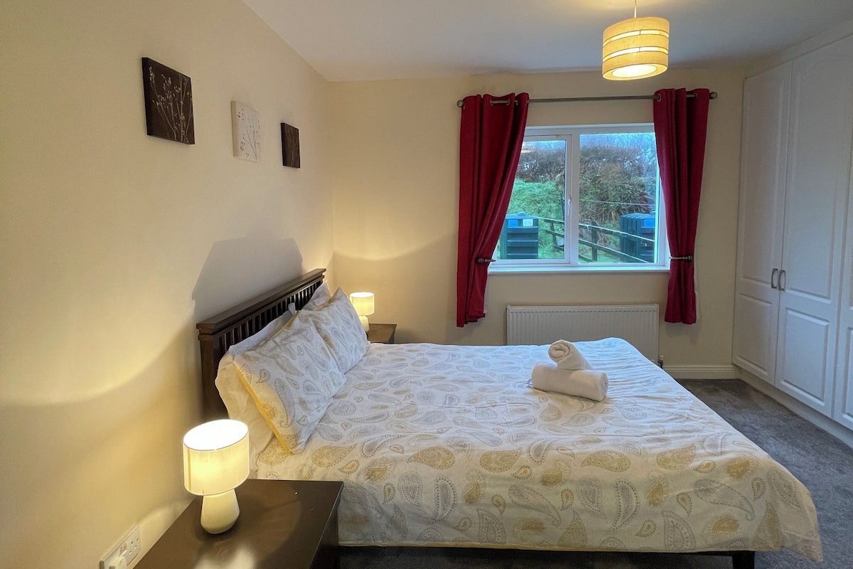 A bedroom with a bed, bedside table, lamp, window with curtains, and a radiator.