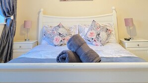 A neatly made bed with a rolled towel, a painting of a pier on the wall, and two bedside lamps. - Valentia Skellig Rooms (Valentia Island)