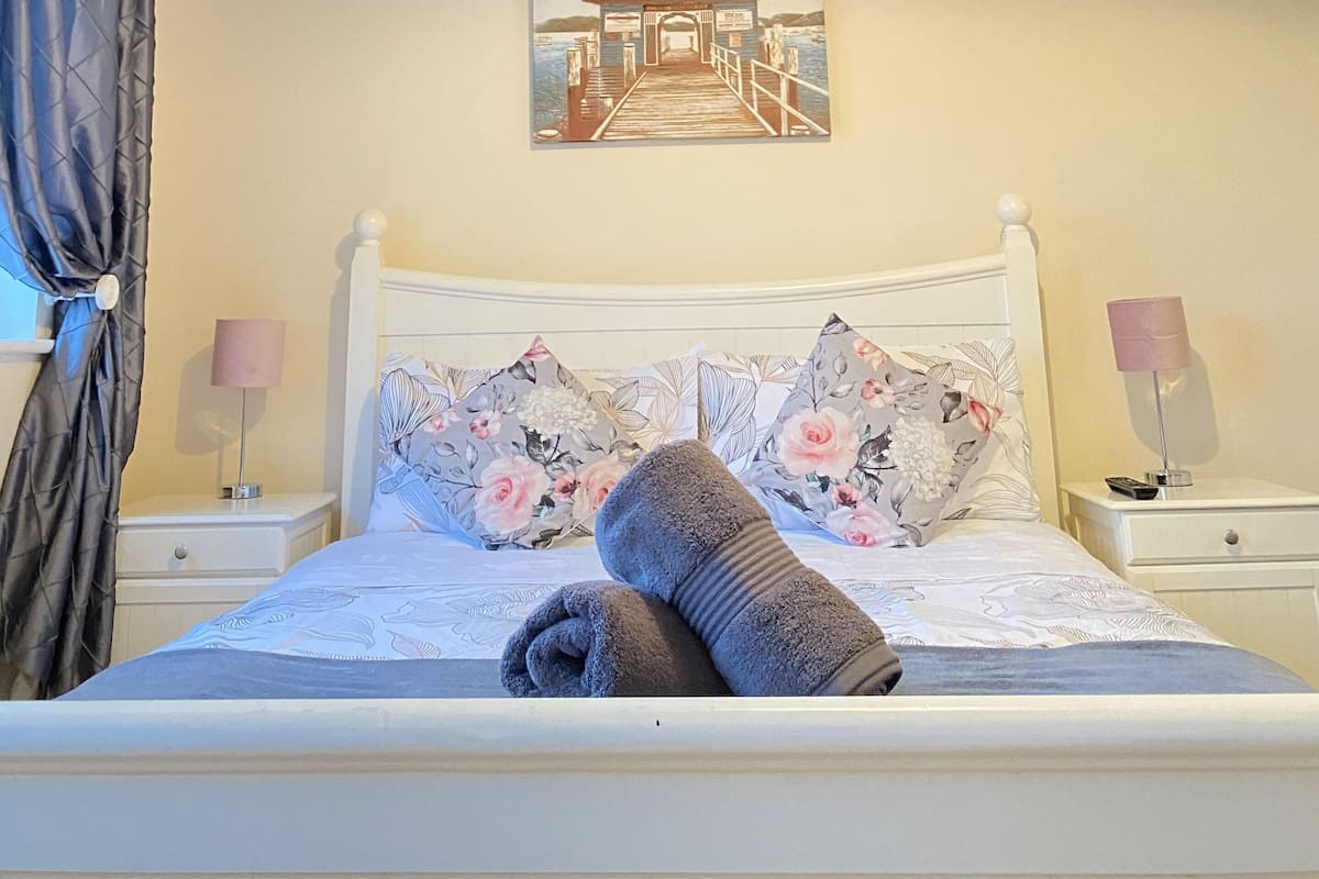 A neatly made bed with a rolled towel, a painting of a pier on the wall, and two bedside lamps.