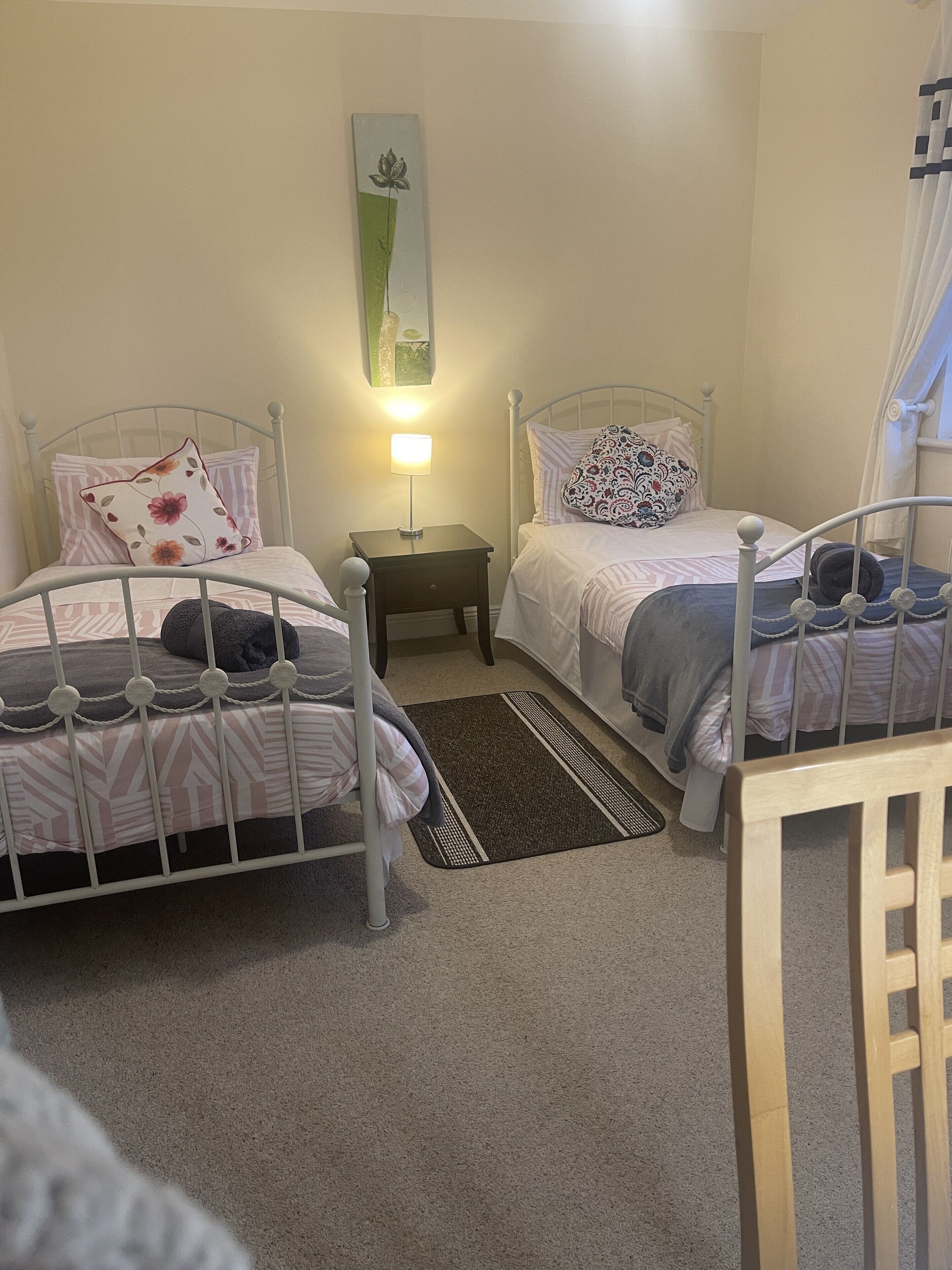A bedroom with two metal beds, a nightstand with a lamp, a small rug, and a painting on the wall.