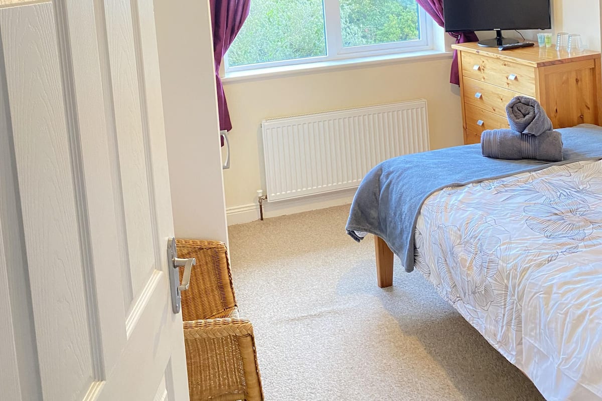 A bedroom with a bed, a television, a radiator, a window with purple curtains, and a door with a number.