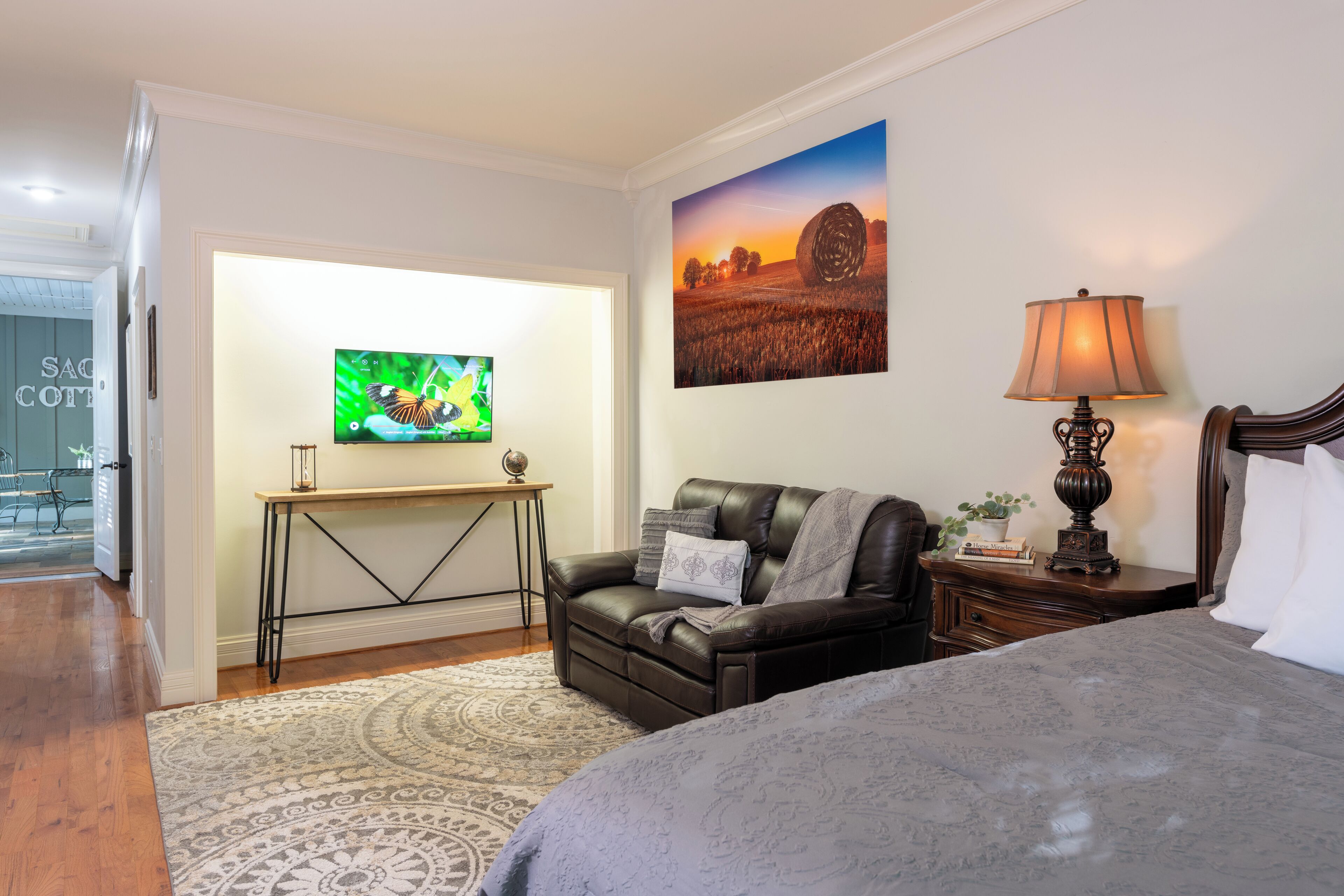 A bedroom with a bed, a desk with a television, a leather sofa, a lamp, and a painting on the wall.
