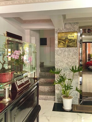 A reception desk with a sign, a glass display case with flowers, a staircase, and a wall with a plaque. - HOANG CAM HOTEL (Ho Chi Minh City)