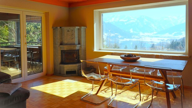 A dining area with a wooden table and clear glass chairs, a stone fireplace, and a large window with a mountain view. - Chalet Lake View (Zell am See)