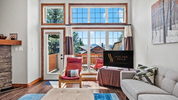 A living room with a fireplace, a red chair, a grey sofa, and a television displaying 'Aisling Baile Property Management'. - Modern Radium Retreat | Fireplace | Mountain Views (Radium Hot Springs)