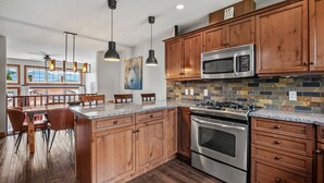 A modern kitchen with wooden cabinets, a stainless steel stove, and a microwave. There is a dining area with a wooden table and chairs. - Modern Radium Retreat | Fireplace | Mountain Views (Radium Hot Springs)