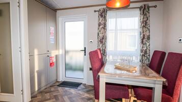 A dining area with a table set for four, red upholstered chairs, a wooden floor, a ceiling light fixture, and a door with a fire extinguisher sign.