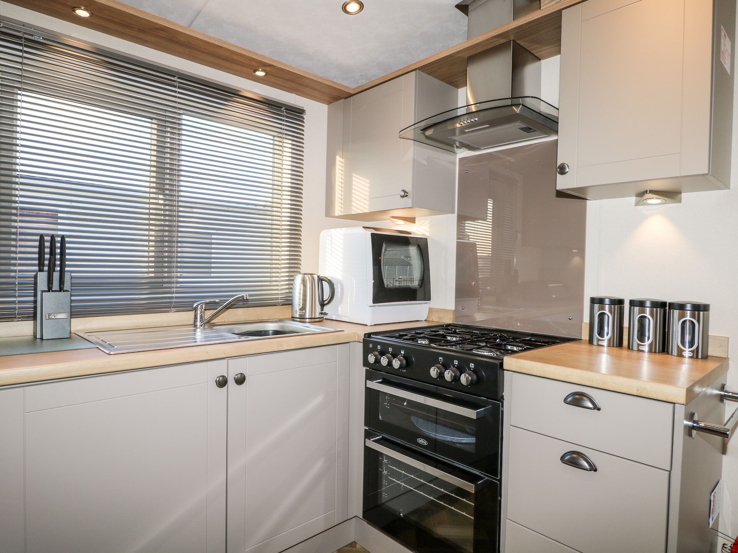 A compact kitchen with a built-in oven, a stovetop, a sink, and a window with blinds.