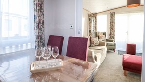 A hotel room with a dining area featuring a wooden table with wine glasses and a tray, a sofa with floral cushions, and a red ottoman.