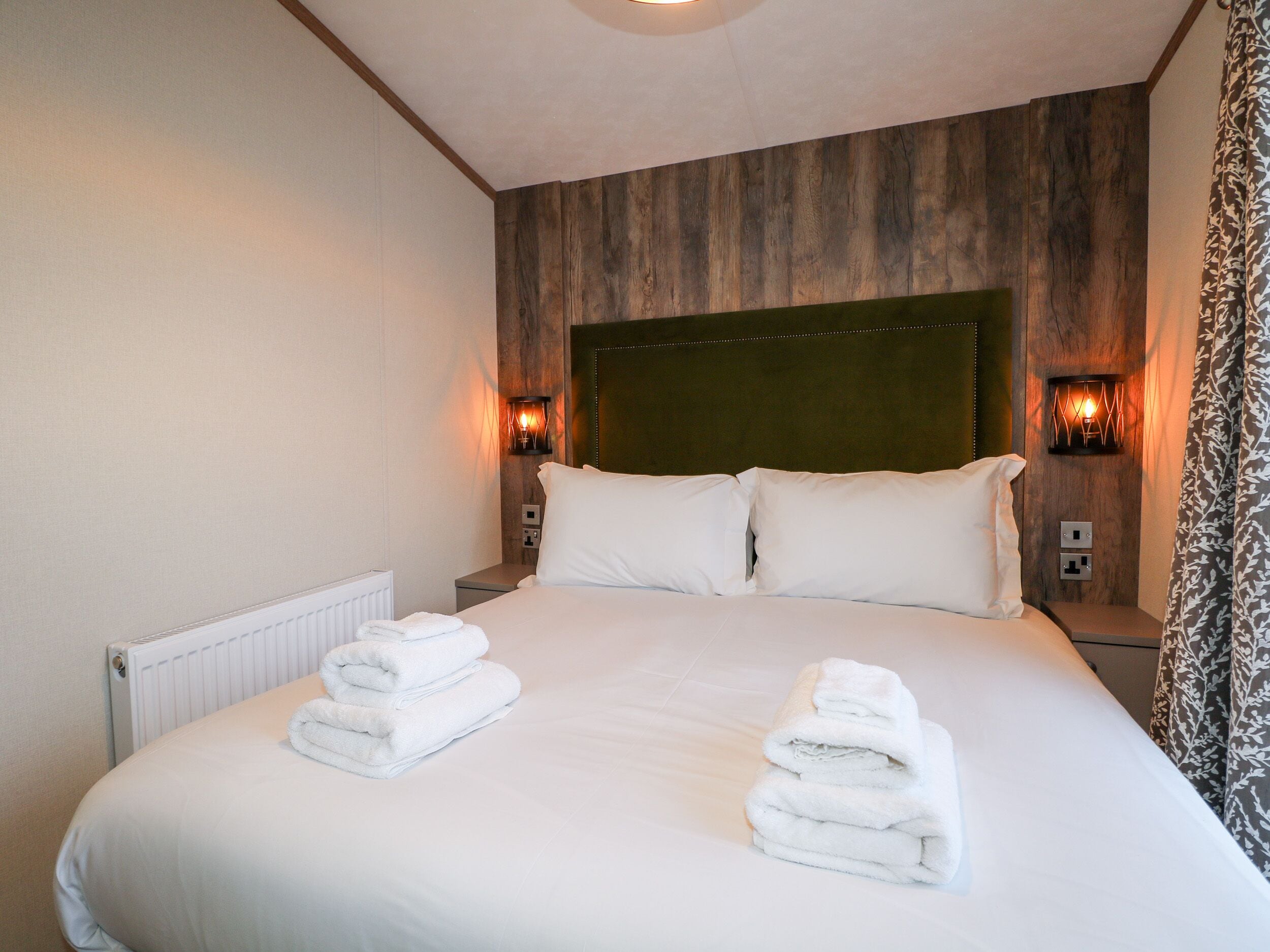 A neatly made bed with white linens and towels, a wooden headboard, and two wall-mounted lamps.