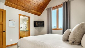 A bedroom with a wooden ceiling, a bed with white bedding, a mounted TV, and a window with curtains.