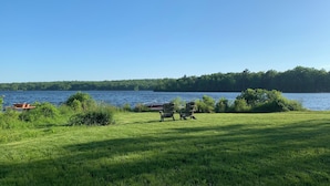 A grassy area with two Adirondack chairs facing a lake, a dock with a boat, and a forested shoreline. - Breathtaking Private, Lakefront Retreat.  (Lebanon)