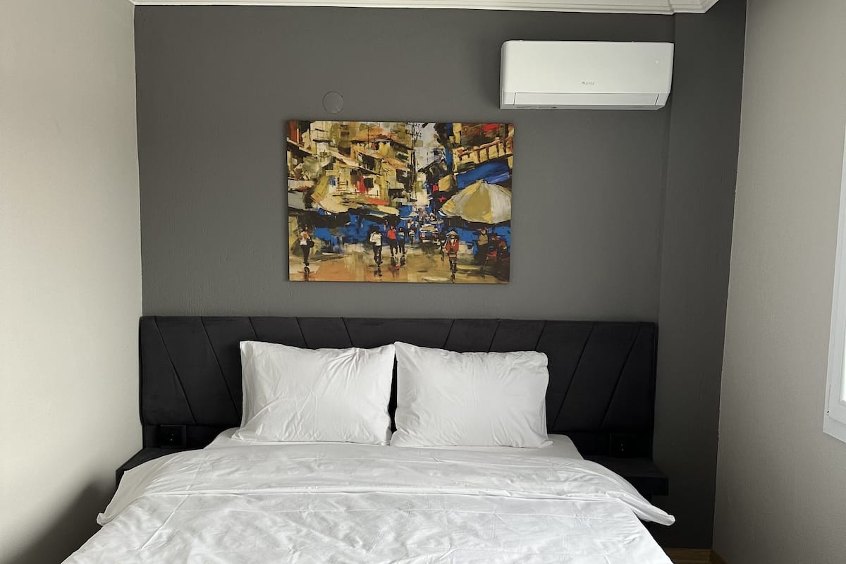 A bedroom with a bed, white bedding, a gray wall, a painting, and an air conditioning unit.