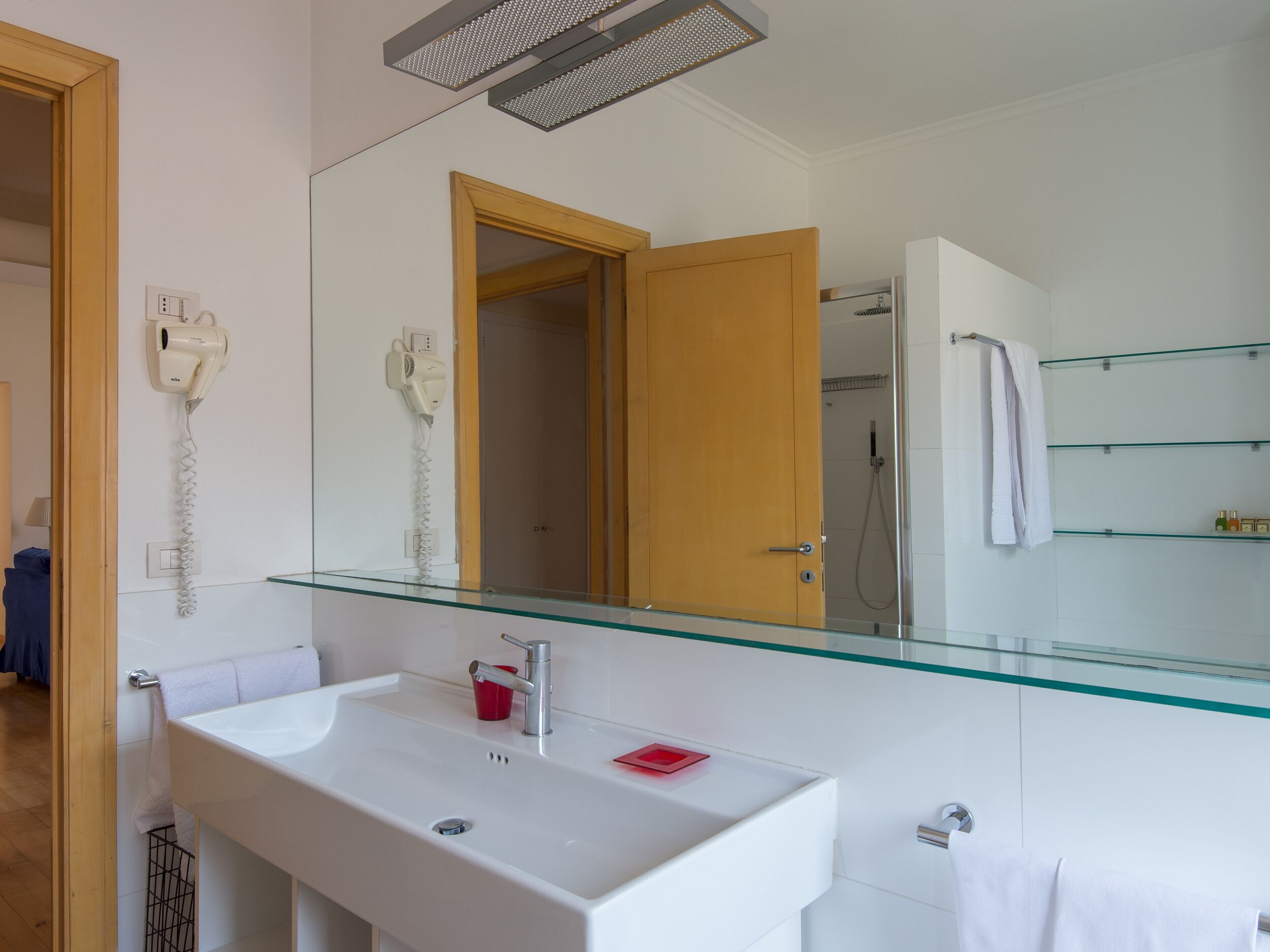 A modern bathroom with a glass shelf, a white sink, a mirror, and a hairdryer mounted on the wall.