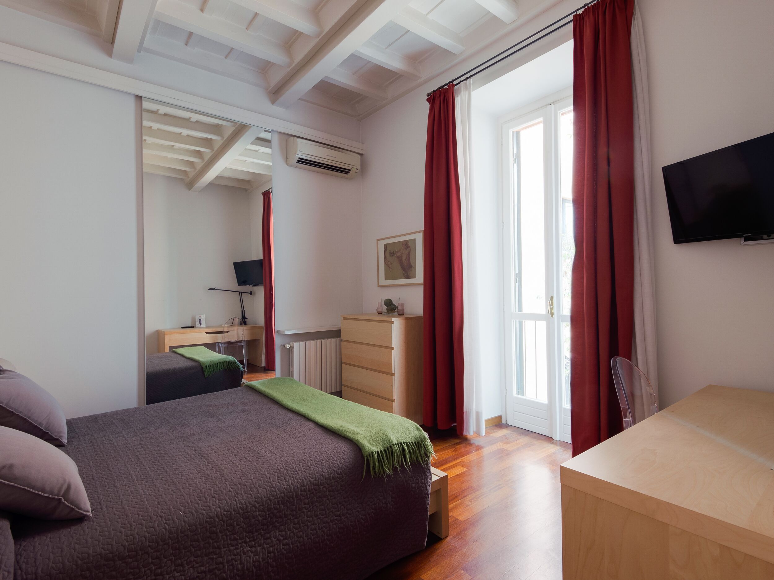 Ein Schlafzimmer mit Bett, Schreibtisch, Kommode, Fernseher und einem Fenster mit Vorhängen.