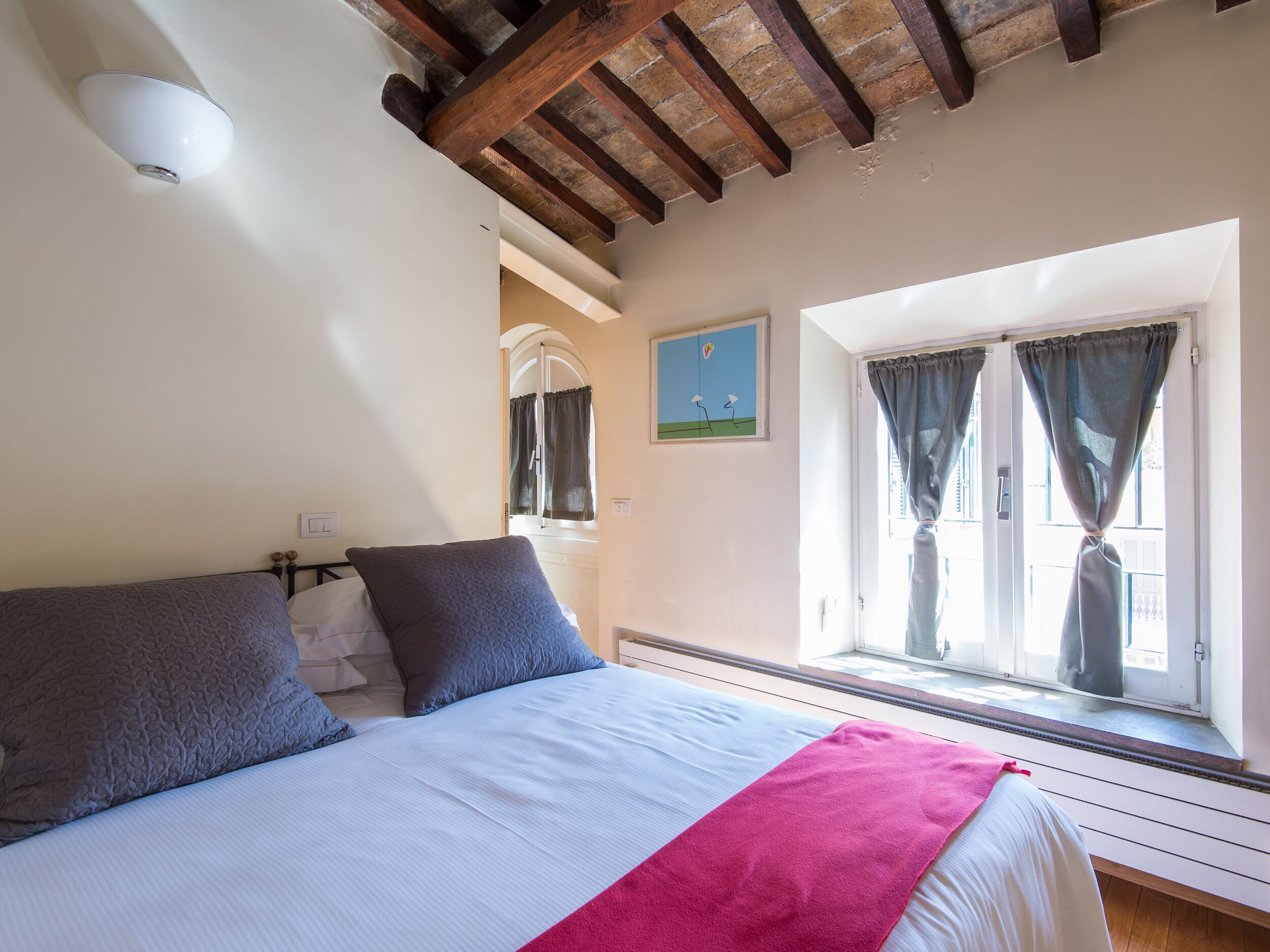 A bedroom with a bed, a window with curtains, a painting on the wall, and a ceiling with exposed beams.