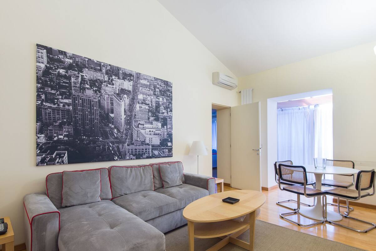A modern living room with a grey sofa, a wooden coffee table, and a dining area with a table and chairs. A large black and white cityscape painting is on the wall.