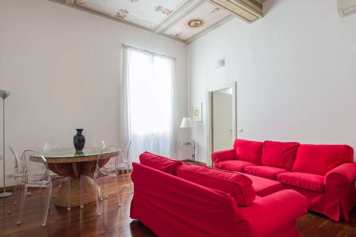 A room with a red sofa, a glass table with chairs, and a large window with curtains.