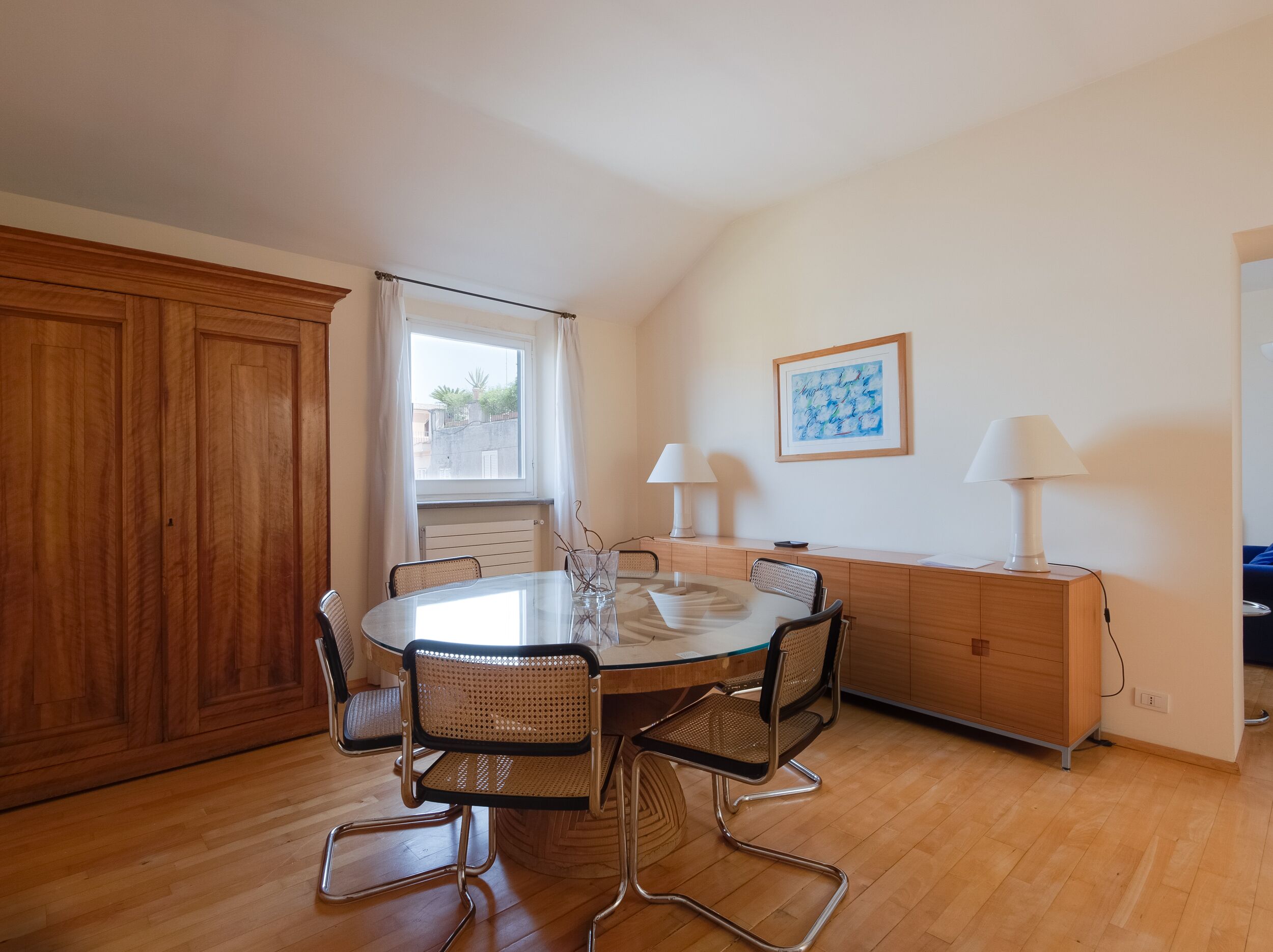 A modern dining area with a round table, four wicker chairs, two white lamps, and a wooden cabinet.