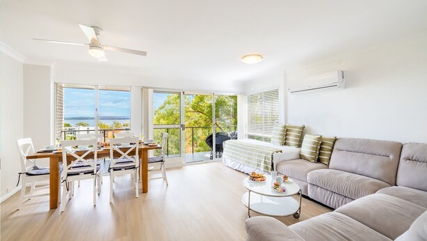 A spacious living room with a large window offering a view of the sea, a ceiling fan, a sofa, and a dining area with chairs and a table. - Sunset Towers 8, 25 Donald St - heart of town with water views (Nelson Bay)
