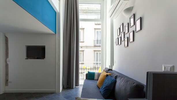 A modern living room with a grey sofa, a flat-screen TV, and a balcony with a view of buildings. - Manuela Apartments San Domenico Centro Storico (Napoli)