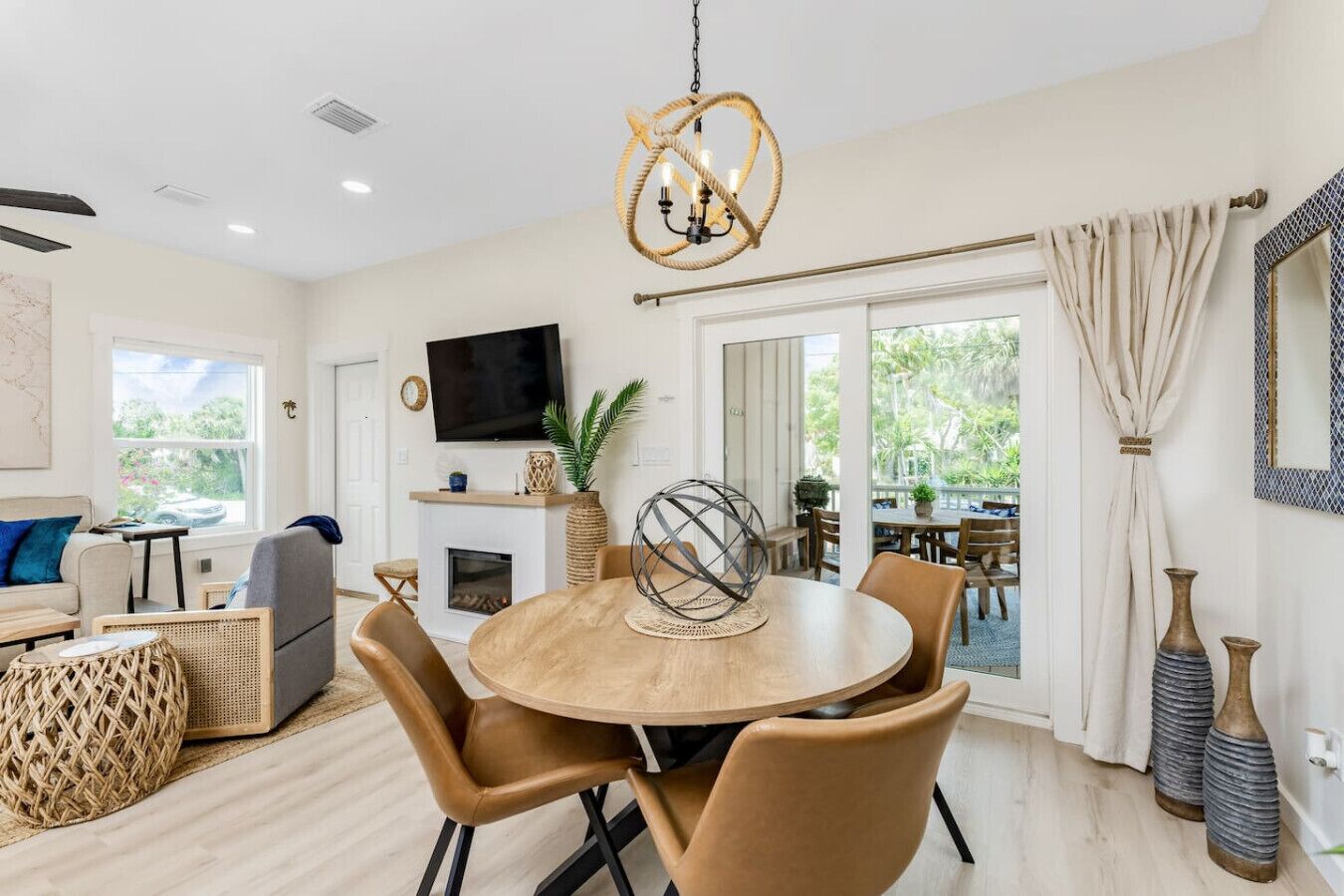 A modern living room with a round wooden table, leather chairs, a flat-screen TV, a fireplace, and a large window with a view of greenery.