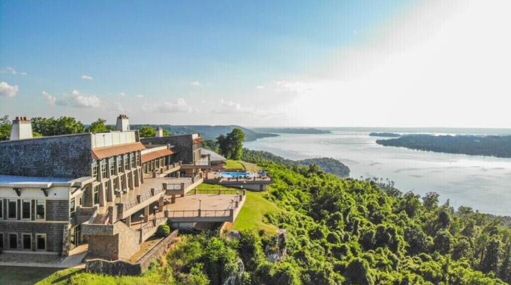 A large building with multiple levels and a swimming pool, overlooking a body of water and lush greenery.