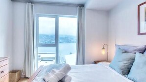 A bedroom with a large window, a bed with white linens, a bedside lamp, and a view of the city. - Serenity by the Slopes: Tremblant Waterfront Condo (Mont-Tremblant)