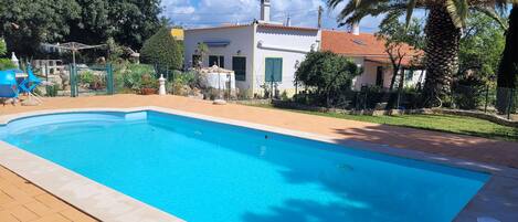 A rectangular swimming pool with a tiled deck, surrounded by a fenced garden with palm trees and a white building with a red roof.