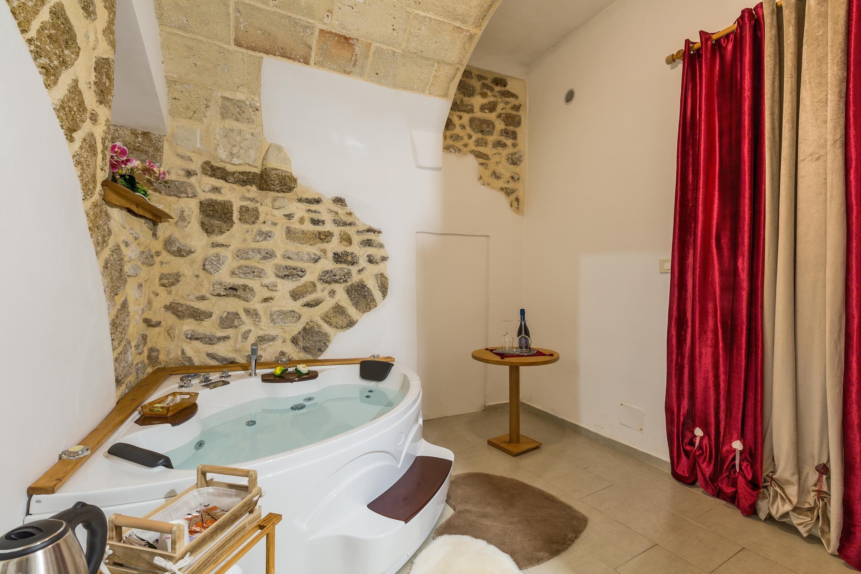 A bathroom with a stone wall, a bathtub, a small table with a bottle, and red curtains.