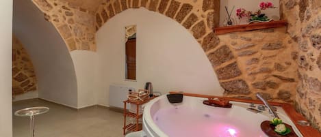 A stone-walled room with a large bathtub, a wooden table with a wine glass, and a shelf with flowers.