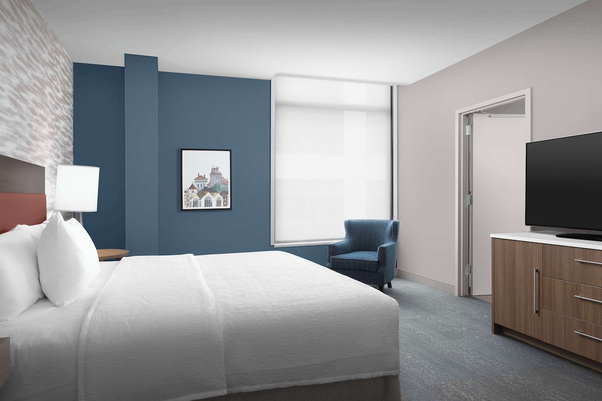 Une chambre d’hôtel avec un grand lit, une chaise, une télévision et une fenêtre avec des stores.