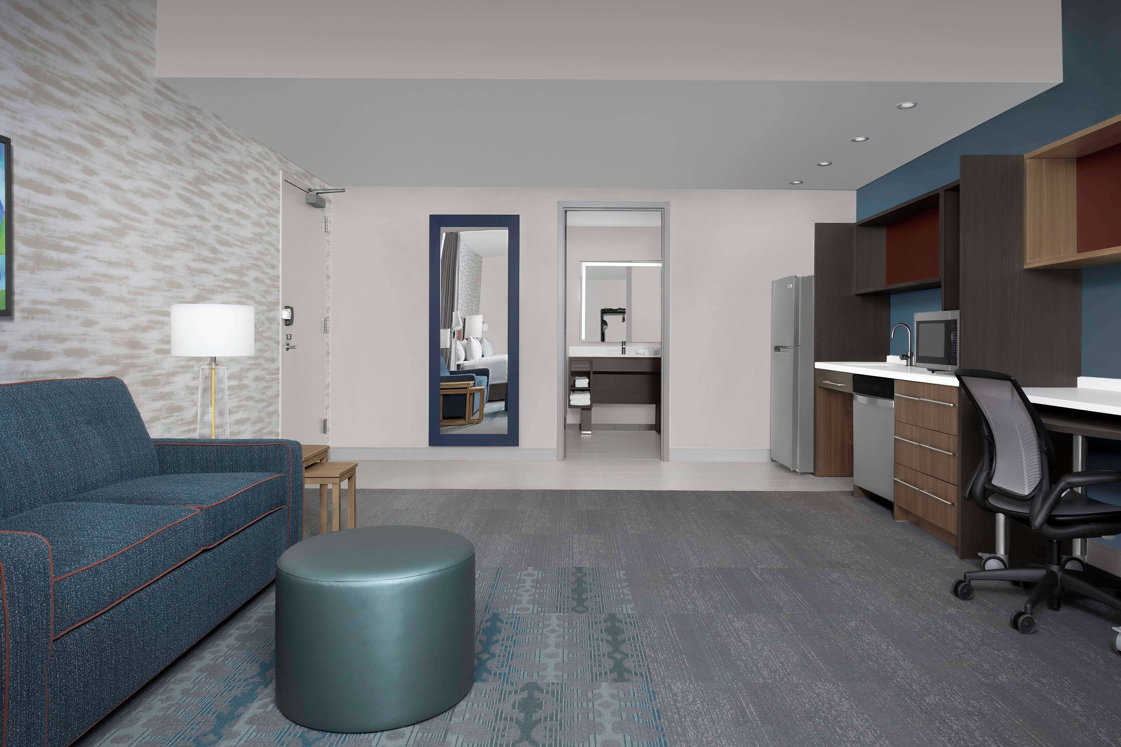 A modern hotel room with a sofa, a round ottoman, a desk with a chair, a kitchenette, and a bathroom visible through a door.