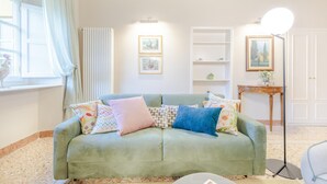 A green sofa with colorful cushions, a white radiator, a shelf with framed pictures, and a floor lamp. - Casa Arnolfo in Lucca (Lucca)