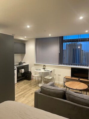 Uma sala de estar moderna com sofá, mesa de jantar com cadeiras, televisão e área de cozinha com armários e janela com persianas.