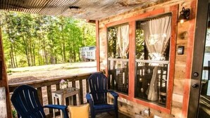 A wooden cabin with a porch, two Adirondack chairs, a small table with a vase, and a view of a house in the background. - Waterfront, 100 Year-Old wood Cabin - #16 (Charlotte)